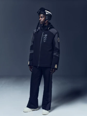 Male model poses in black BOSS ski jacket, ski pants, helmet, and snow boots against a grey gradient backdrop in a studio shot.