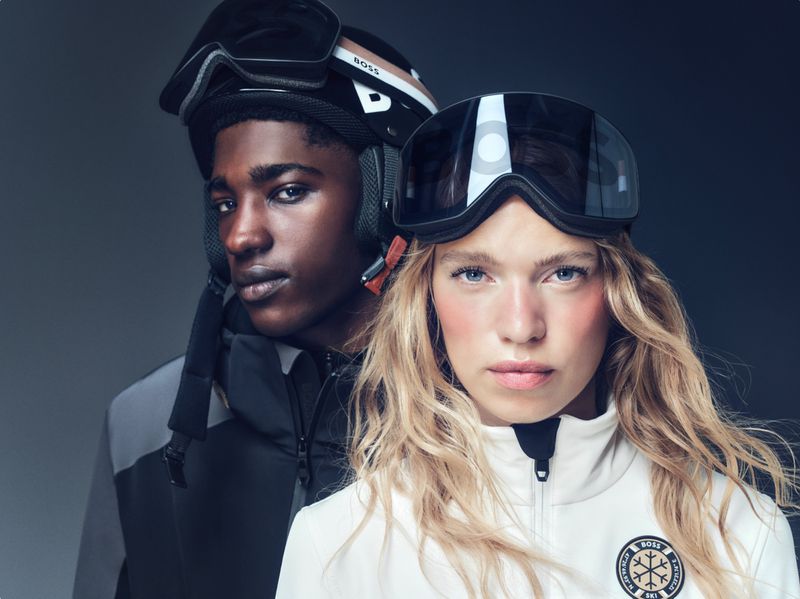 Two fashion models pose in ski apparel and helmets from BOSS. One is a blond woman wearing goggles and the other is a Black man.