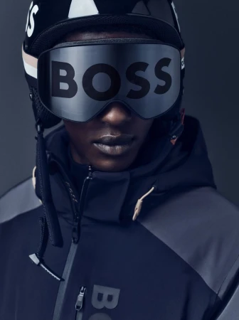 Close-up of a black adult male wearing a black helmet and goggles that read 'BOSS', in a dark studio. He is wearing a dark ski jacket.
