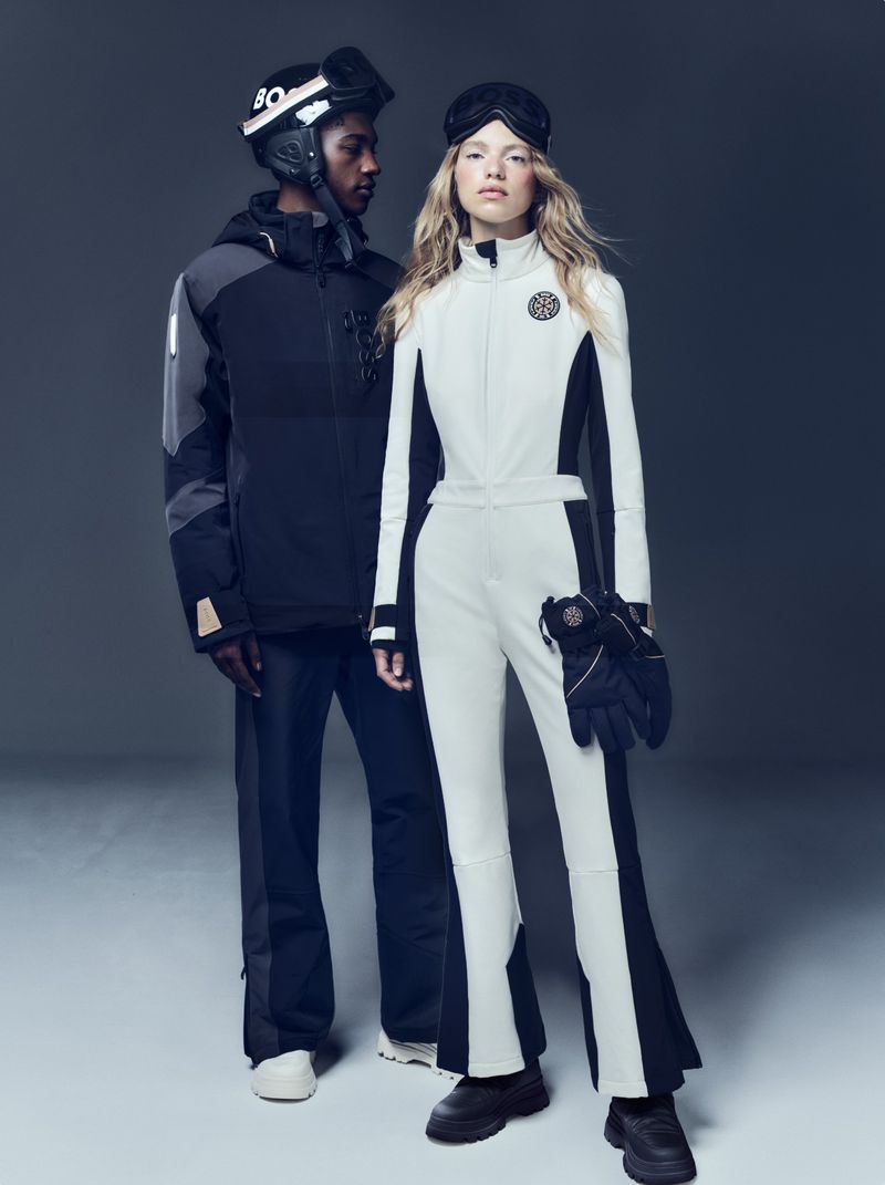 Two people in winter sports clothing in a studio setting. One male in black and gray and one female in white. Dark, neutral background.