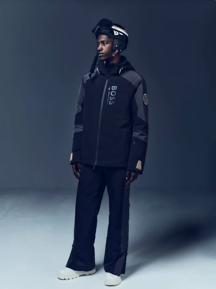 Model in black ski jacket and matching pants wearing a ski helmet against a neutral gradient background