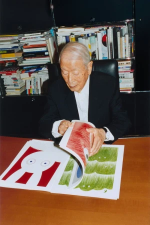 Elderly Japanese man seated, flipping pages of a children's illustration book, surrounded by a bookshelf and dark wood desk.