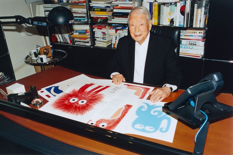 Elderly man in a black suit sits at a desk with colorful cartoon drawings, books, and a lamp in an indoor setting with bright lighting.