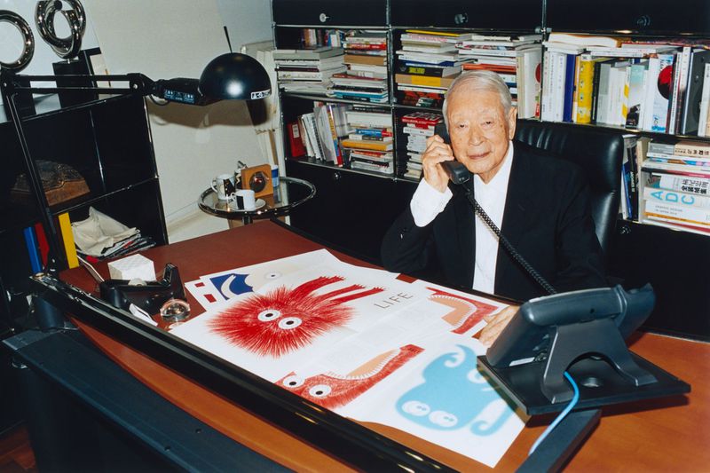 Elderly man in a suit sits at his desk while on the phone, surrounded by books, documents and artwork in a well-lit office.