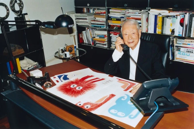 Elderly man in a suit sits at his desk while on the phone, surrounded by books, documents and artwork in a well-lit office.