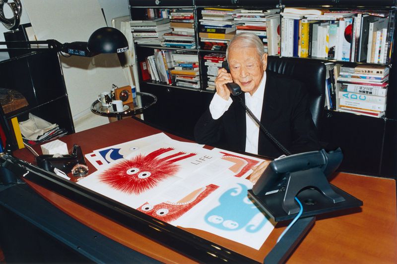 Elderly Asian man in a suit sits at a desk, talking on the phone with cartoon artwork and bookshelves behind him.