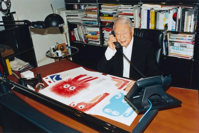 Elderly Asian man in a suit sits at a desk, talking on the phone with cartoon artwork and bookshelves behind him.