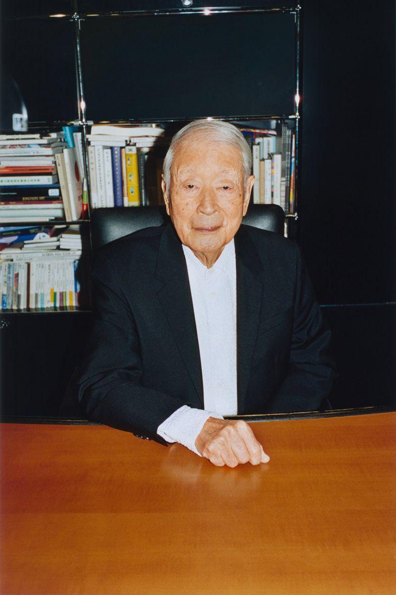 Japanese graphic designer Kazumasa Nagai seated at a wooden desk wearing a black blazer and white shirt in front of bookshelves.