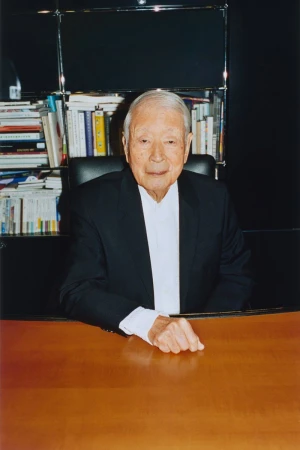 Japanese graphic designer Kazumasa Nagai seated at a wooden desk wearing a black blazer and white shirt in front of bookshelves.