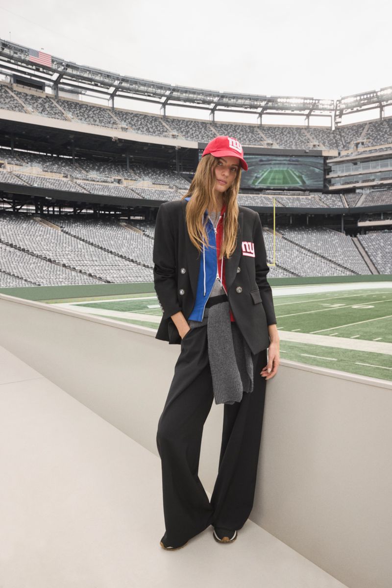 Fashion model poses in a stadium wearing a black blazer, wide-leg pants, and a red baseball cap with stadium seating in the background.