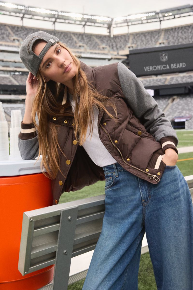 A woman wearing a brown jacket, jeans, and a baseball cap poses at an outdoor stadium, leaning against an orange and gray bench.