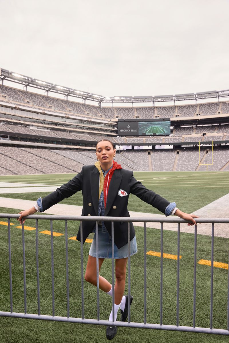 A young adult woman poses confidently in front of a football stadium, wearing a blazer, skirt, socks, and loafers.