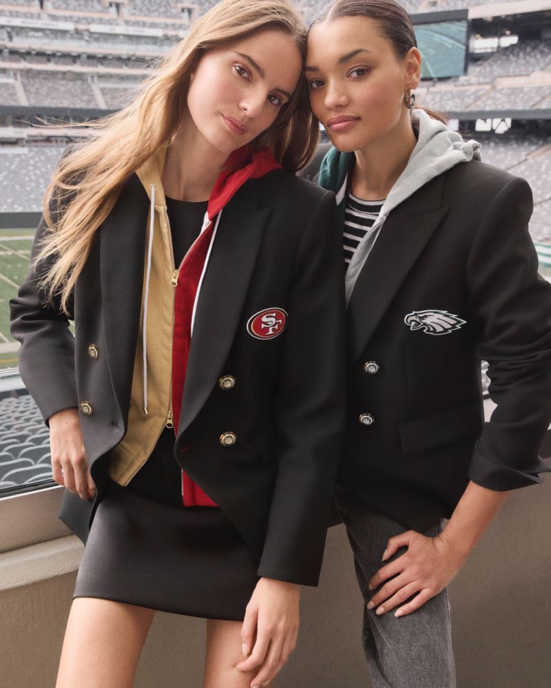 Two women posing in football jackets with stadium background. Fashionable outfits. San Francisco 49ers and Philadelphia Eagles attire.