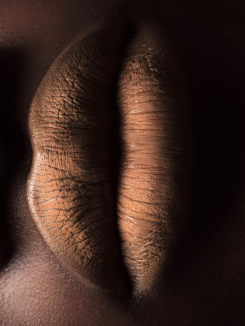 Close-up of an adult African American woman's tan lips, set against a contrasting dark background emphasizing their texture and form.