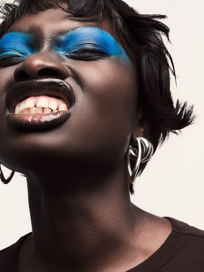 Close-up studio shot of a Black woman with blue eyeshadow and glossy lips wearing silver earrings against a neutral backdrop.