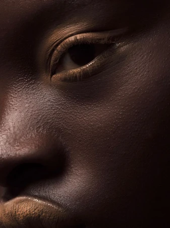 Close-up portrait of an adult female with brown skin in a studio setting, focusing on the skin's texture and the model's eye.