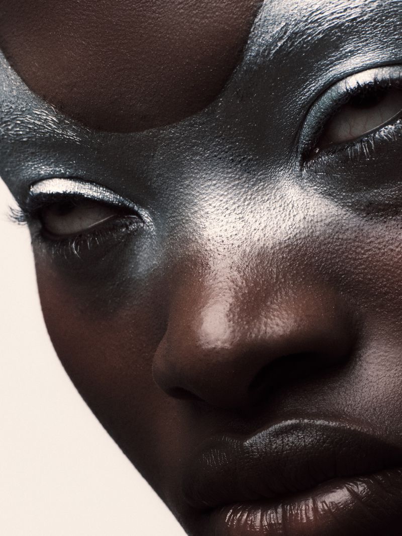 Close-up of a dark-skinned female with dramatic silver face paint, contrasting with white accents around the eyes and glossy lips.