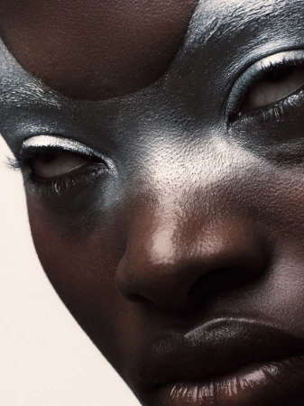 Close-up of a dark-skinned female with dramatic silver face paint, contrasting with white accents around the eyes and glossy lips.