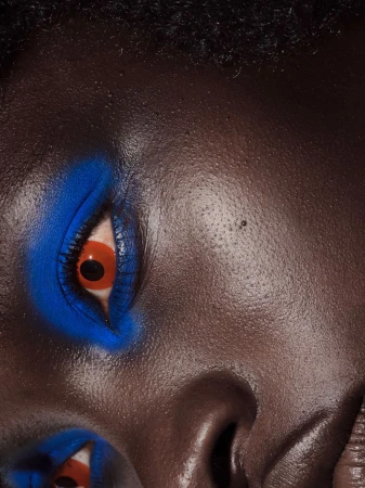 Close-up of a dark-skinned person's face featuring bold blue eye makeup and striking orange-red eye contacts, showcasing dramatic beauty style.