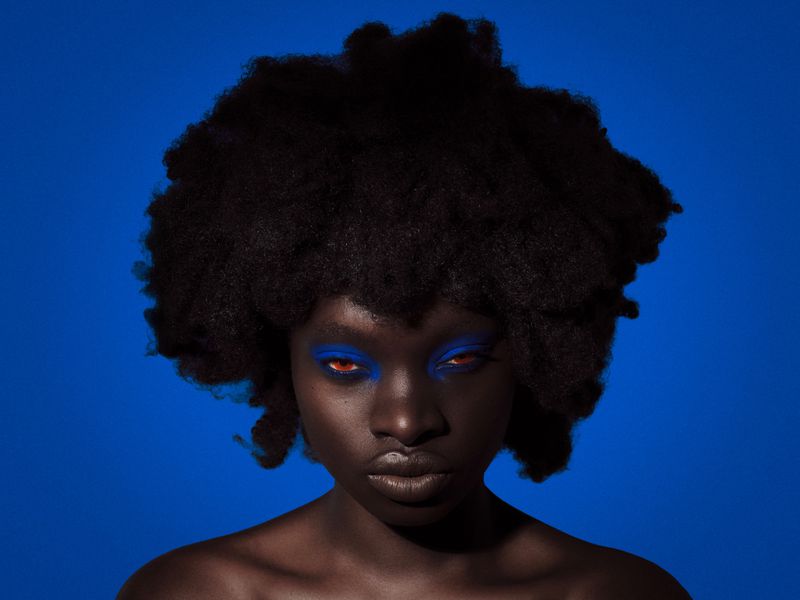 Black woman with a voluminous afro and vibrant blue eye makeup posed against a solid royal blue background.