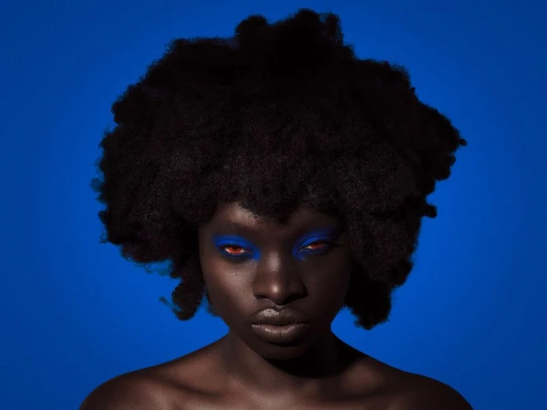 Black woman with a voluminous afro and vibrant blue eye makeup posed against a solid royal blue background.
