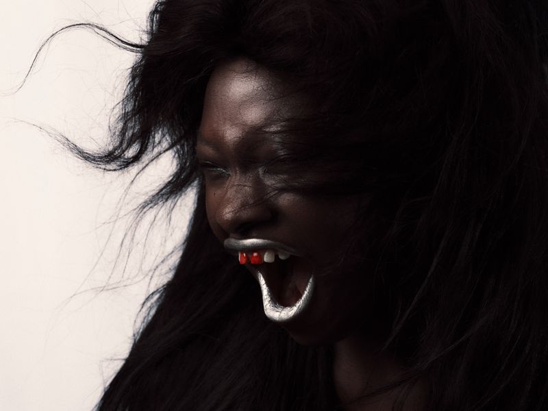 Portrait of a person with dark skin and silver-painted teeth and red tongue, against a light background with visible black wavy hair.