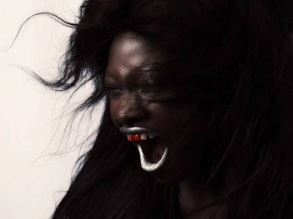 Portrait of a person with dark skin and silver-painted teeth and red tongue, against a light background with visible black wavy hair.