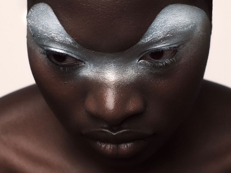 Close-up portrait of a Black woman with dramatic silver eye makeup, looking down with focus, set against a soft, diffused background.