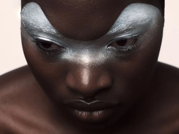 Close-up portrait of a Black woman with dramatic silver eye makeup, looking down with focus, set against a soft, diffused background.