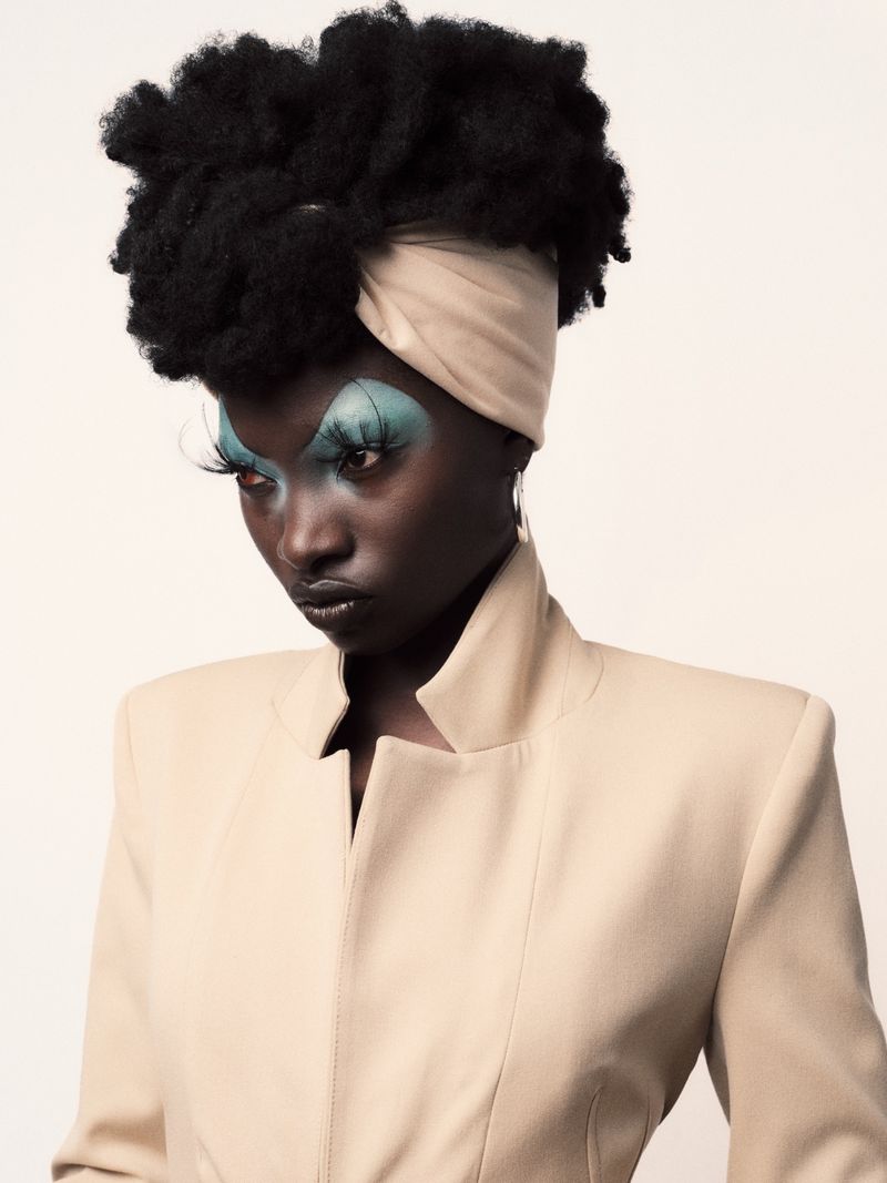 Close-up fashion portrait of a Black woman with afro, wearing a tan jacket and headwrap, against a white backdrop in a photo studio.