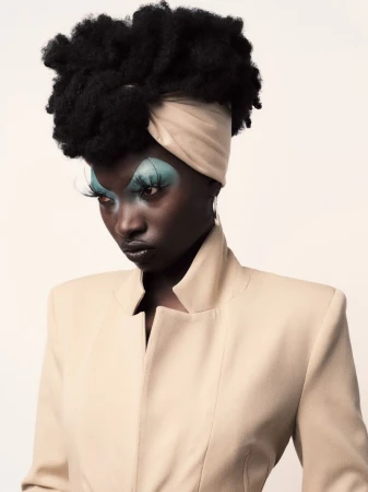 Close-up fashion portrait of a Black woman with afro, wearing a tan jacket and headwrap, against a white backdrop in a photo studio.