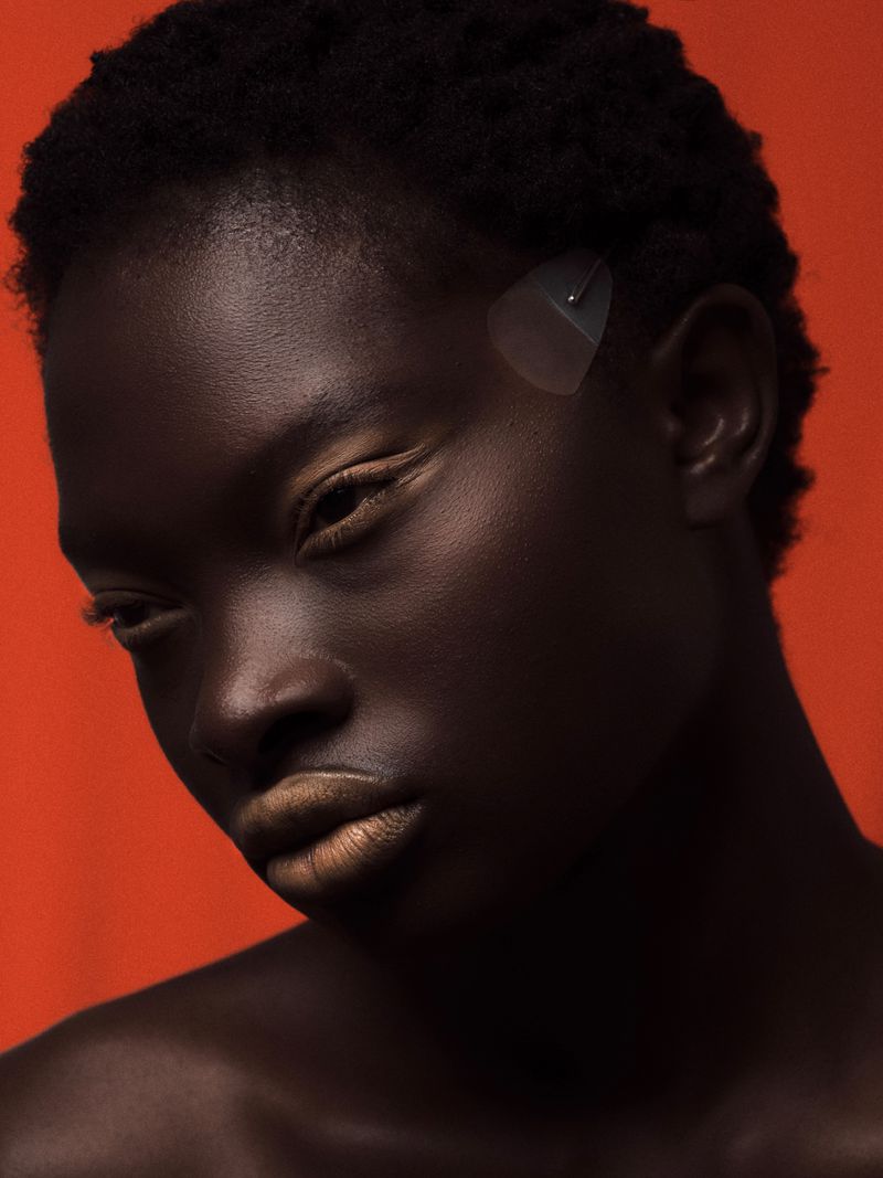 Close-up portrait of a dark-skinned adult female with golden lips and a geometric transparent hair clip against an orange backdrop.
