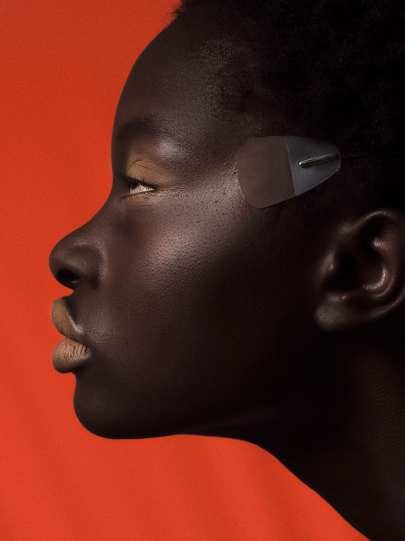 Close-up profile of a woman with dark skin and a sleek hairstyle, wearing a futuristic accessory. The backdrop is a vibrant orange.