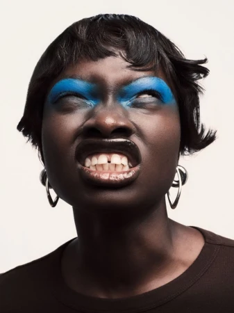 Close-up of a Black woman with bright blue eyeshadow and glossy lips grimacing against a white background.