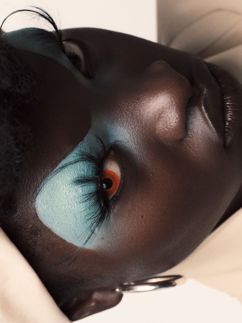 Close-up of a Black woman with light blue eye makeup, long eyelashes, and an orange contact lens, wearing a light-colored head covering.