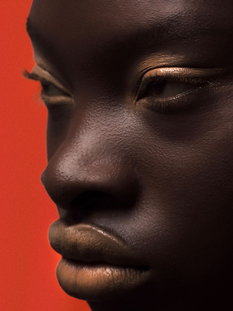Close-up portrait of a dark-skinned adult female with golden eyeshadow and lipstick against a vibrant orange backdrop in a beauty context.
