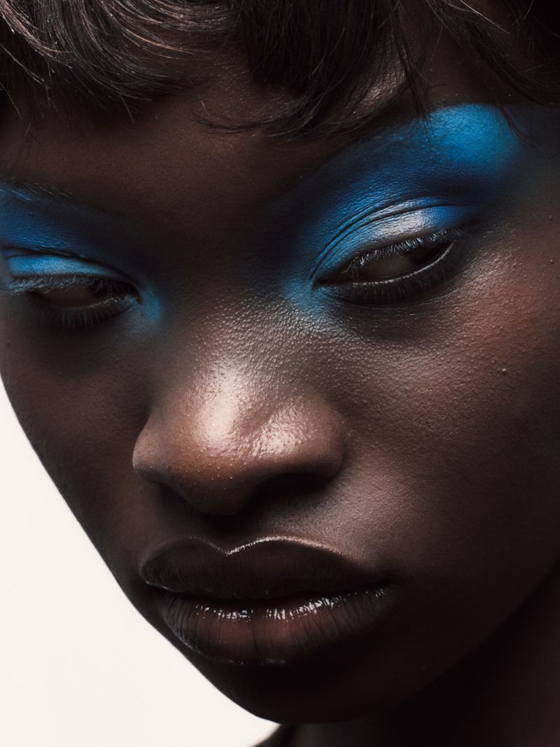 Close-up portrait of a Black woman with glossy lips and bright blue eyeshadow. Beauty shot against an off-white backdrop.