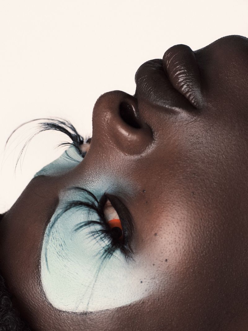 Close-up shot of an African American woman's face with dramatic turquoise and white eye makeup, upward gaze, and flawless dark skin.
