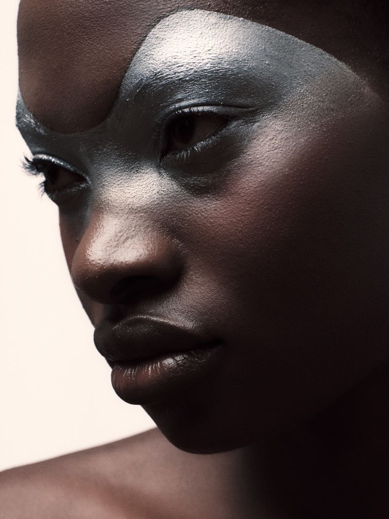 Close-up portrait of an African American female model with silver makeup on her face looking down in a dimly lit photo studio.
