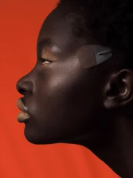 Profile portrait of a Black woman with a translucent eye patch against a solid orange background for Schön Magazine.