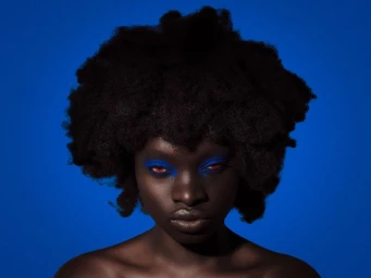 Black woman with a voluminous afro and vibrant blue eye makeup posed against a solid royal blue background.