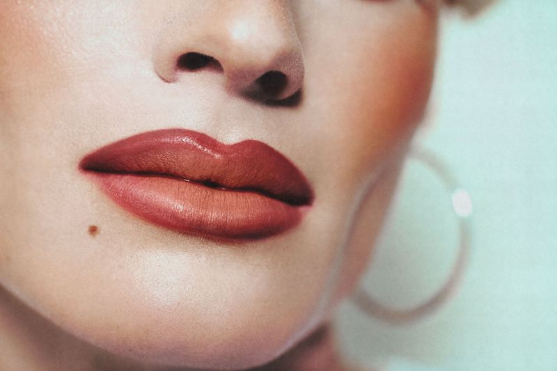 Close-up portrait of an adult female with red lipstick, a mole, and hoop earrings on a white background with soft lighting.
