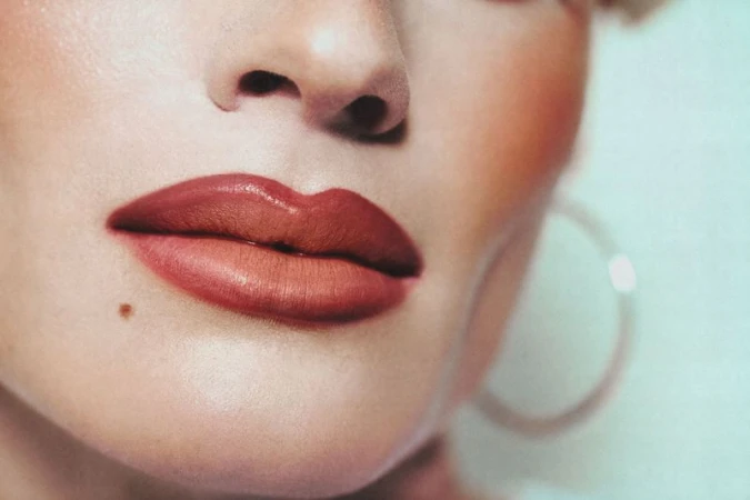 Close-up portrait of an adult female with red lipstick, a mole, and hoop earrings on a white background with soft lighting.