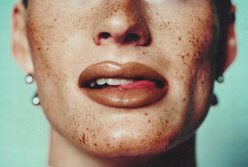 Close-up of a freckled face with glossy brown lips and tongue touching upper teeth against a pale turquoise background.