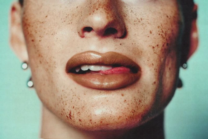 Close-up of a freckled face with glossy brown lips and tongue touching upper teeth against a pale turquoise background.
