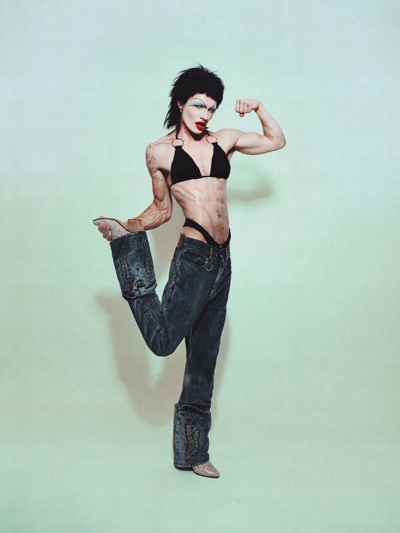 Person posing in jeans, bikini top, and heels, flexing bicep against a pale green background in a studio shot. Fashion concept.