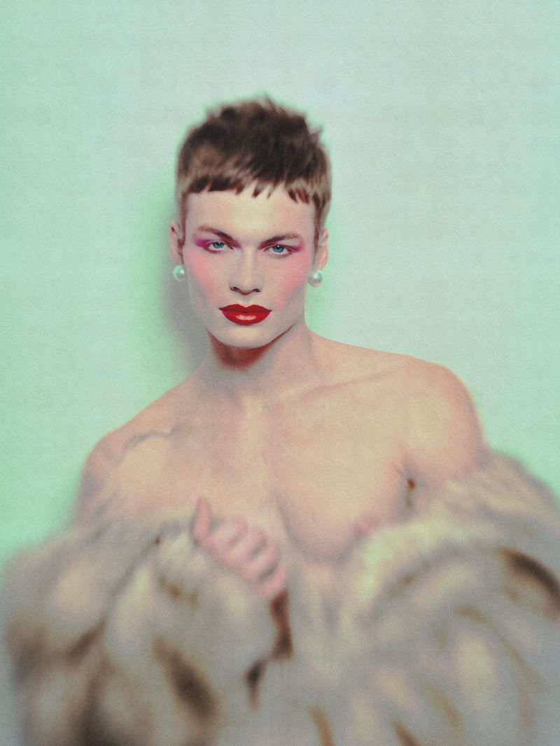 Portrait of a shirtless male model wearing red lipstick, earrings, and blush, partially covered with a blurred fur coat, on a light green background.