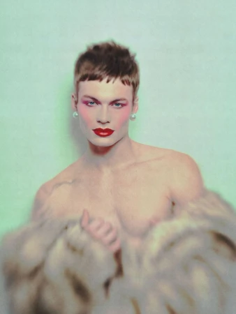 Portrait of a shirtless male model wearing red lipstick, earrings, and blush, partially covered with a blurred fur coat, on a light green background.