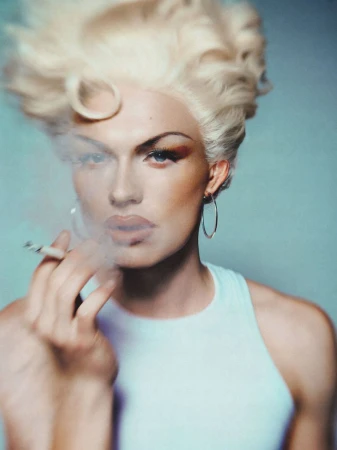 Portrait of a blonde model with sculpted voluminous hair smoking a cigarette in a white top against a soft blue backdrop.