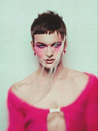 Portrait of a person with short brown hair and pink makeup wearing a pink fuzzy jacket against a light green background.
